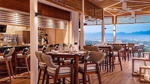A restaurant with wooden furniture, a bar area, and a view of the city and mountains. - Stunning Grand Luxxe 2-Bedroom Loft, Perfect for you to indulge in paradise. (Nuevo Vallarta)