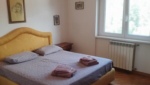 Un dormitorio con una cama, dos almohadas, una mesita de noche, una ventana y un radiador.