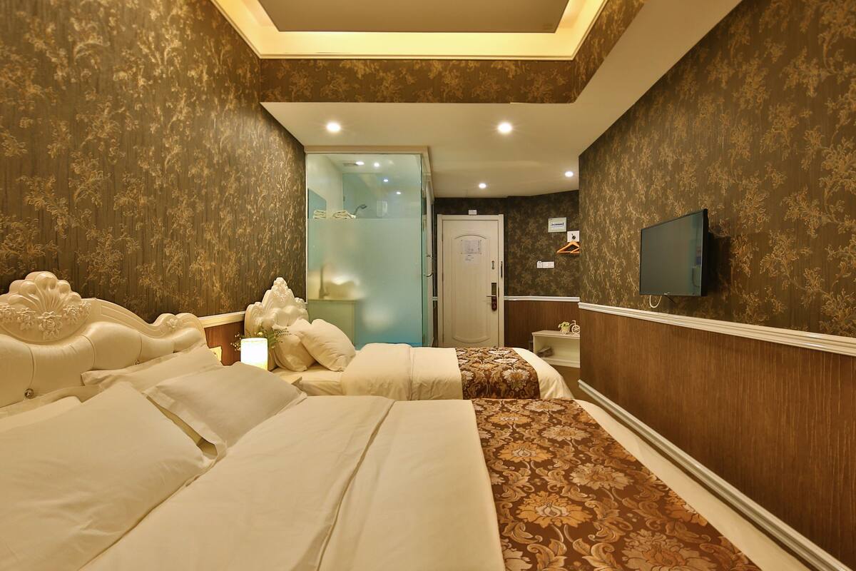 A hotel room with two beds, a shower area, a TV mounted on the wall, and a door leading to another room.