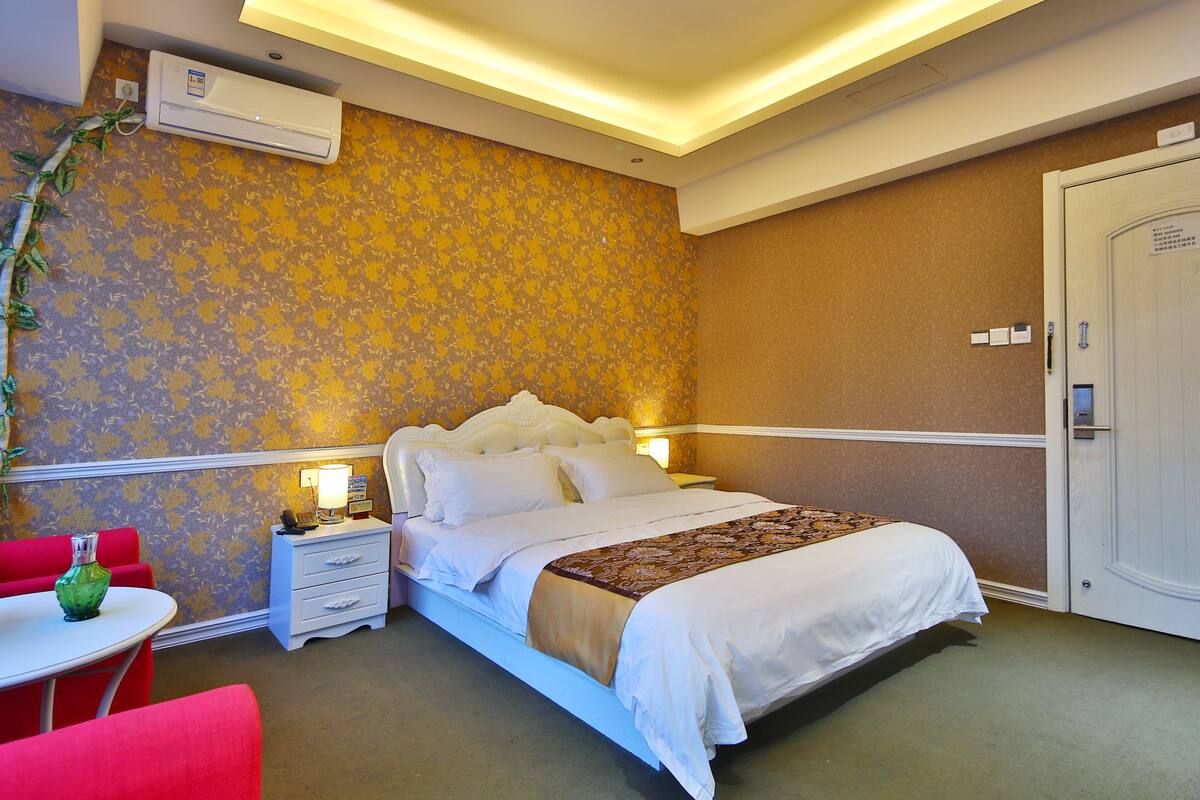 A hotel room with a bed, bedside table, and a small table with a red chair.