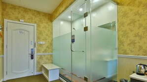 Une pièce avec une douche en verre, une porte blanche, un petit banc et une poubelle.