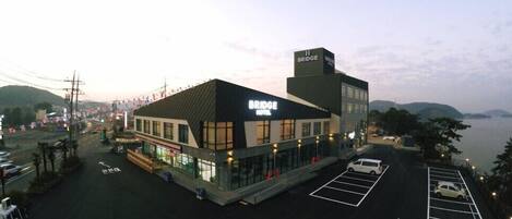 A hotel named 'Bridge Hotel' with a modern design, a parking lot, and a view of the water and hills in the background.