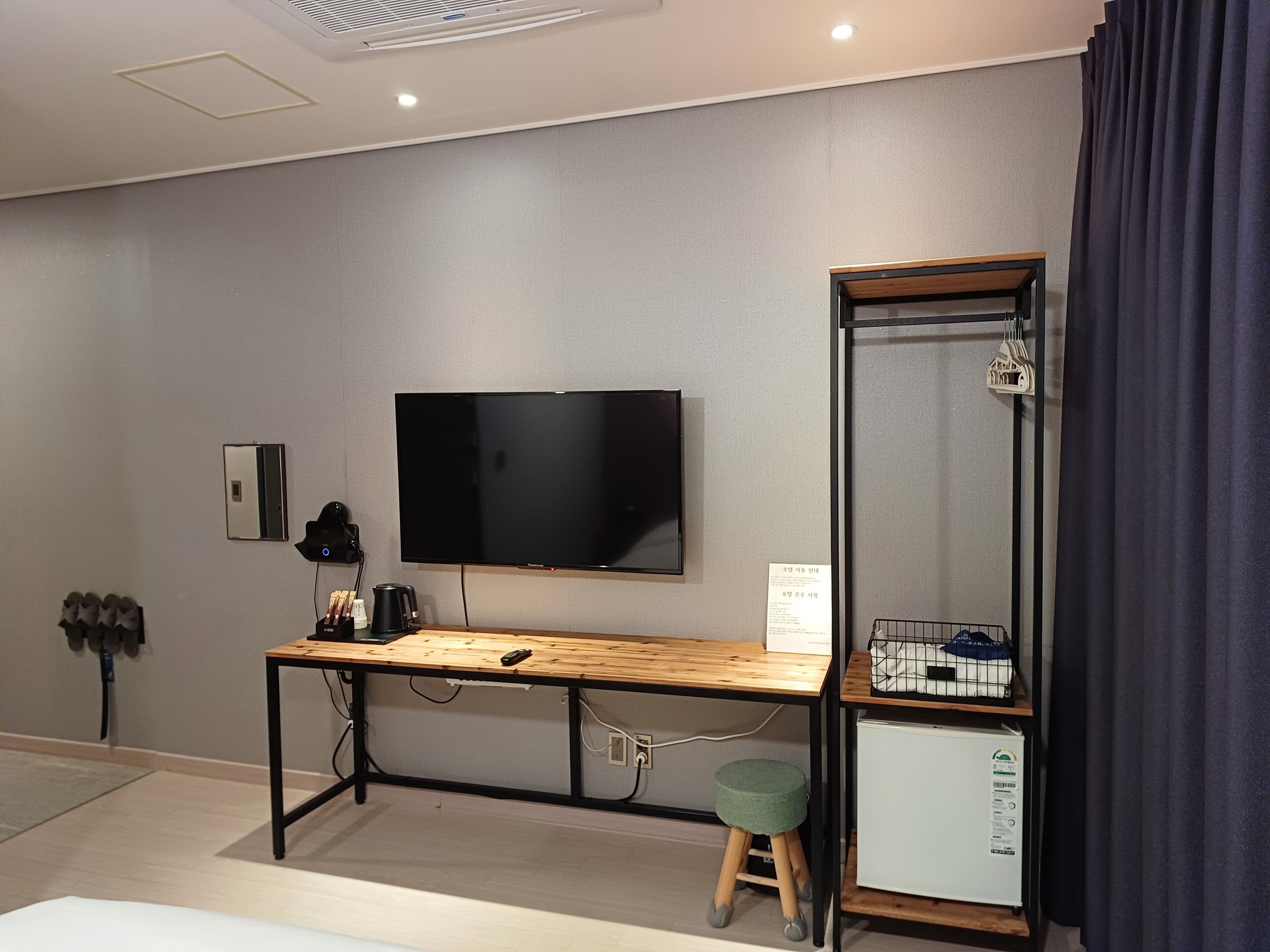 Une chambre d’hôtel moderne équipée d’un téléviseur à écran plat fixé au mur, d’un bureau en bois avec une chaise, d’un petit tabouret, d’un mini-réfrigérateur et d’un miroir.