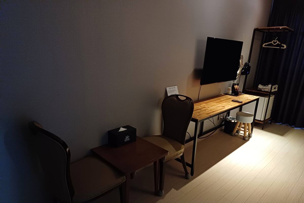 Ein kleiner, schwach beleuchteter Raum mit einem Holztisch, zwei Stühlen, einem an der Wand montierten Fernseher und einem kleinen Tisch mit einer Lampe.