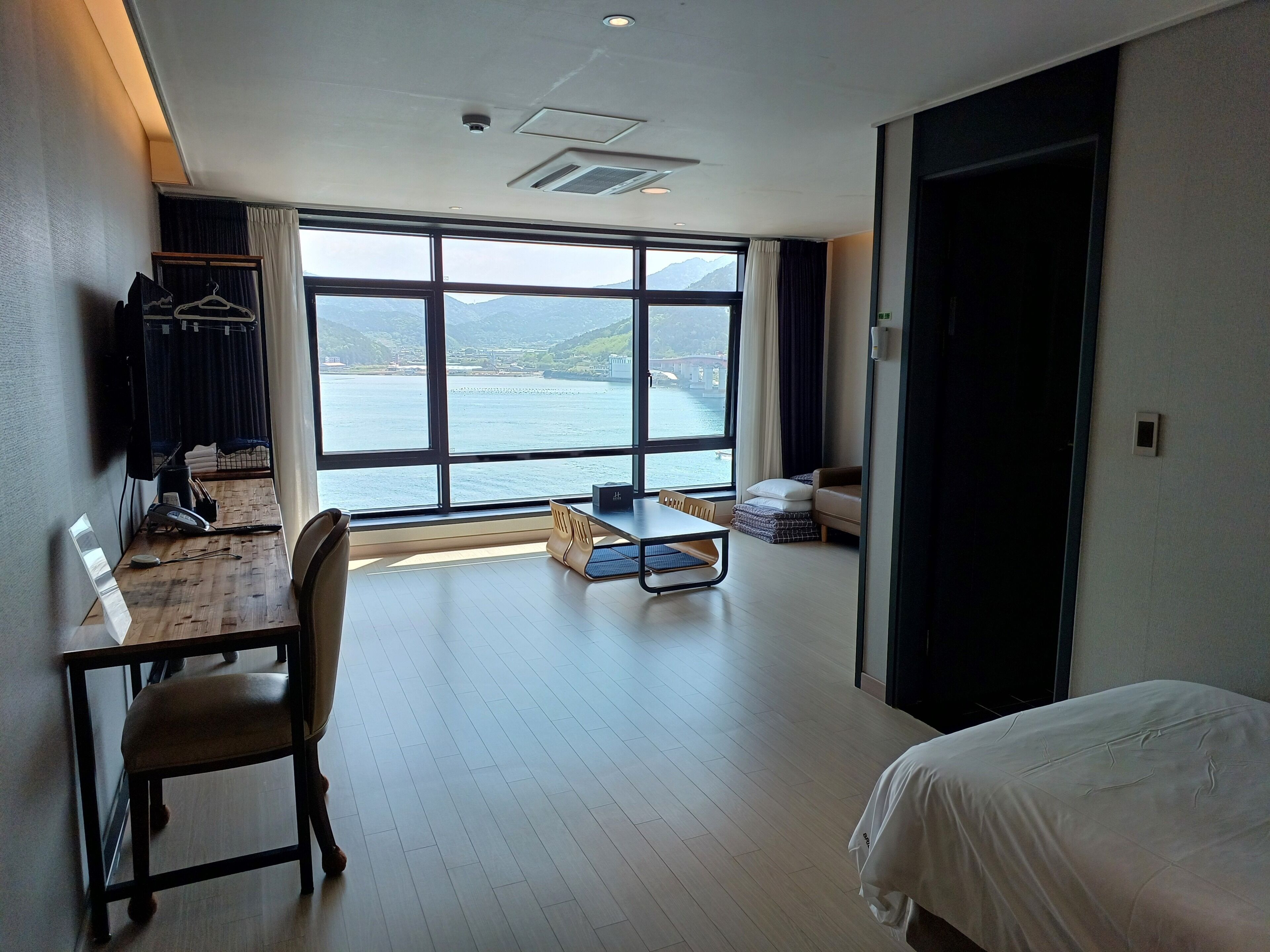 A hotel room with a large window offering a view of the sea and mountains. There is a desk with a chair, a bed, and a small sofa.