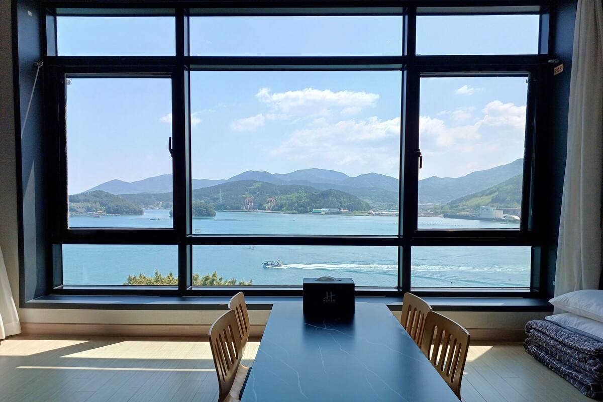 Ein Zimmer mit großem Fenster und Blick auf See und Berge. Ein Tisch mit Stühlen und ein kleines schwarzes Objekt stehen darauf.