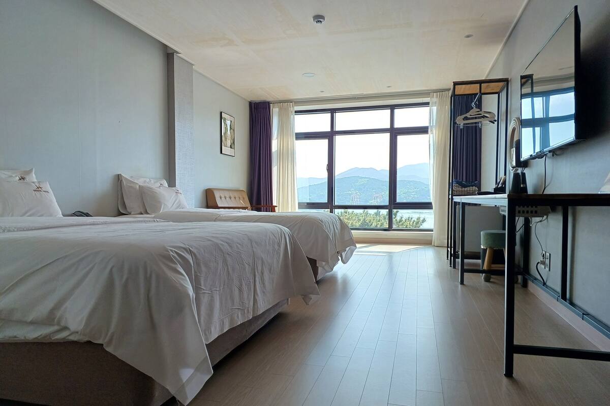 Ein Hotelzimmer mit zwei Betten, einem großen Fenster mit Bergblick, einem Schreibtisch mit Stuhl und einem an der Wand befestigten Flachbildfernseher.