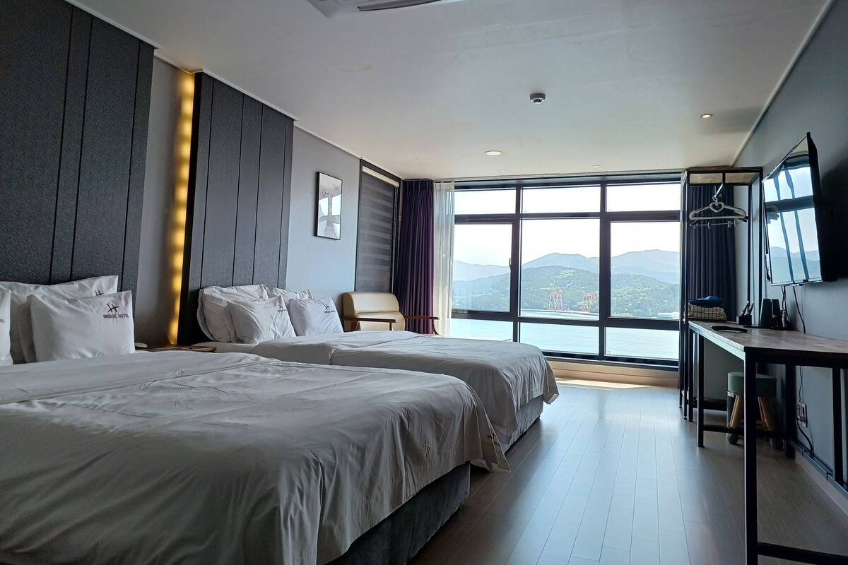 Ein Hotelzimmer mit zwei Betten, einem Schreibtisch, einem Sessel und einem großen Fenster mit Blick auf die Berge, sowie einem Wand-Fernseher.