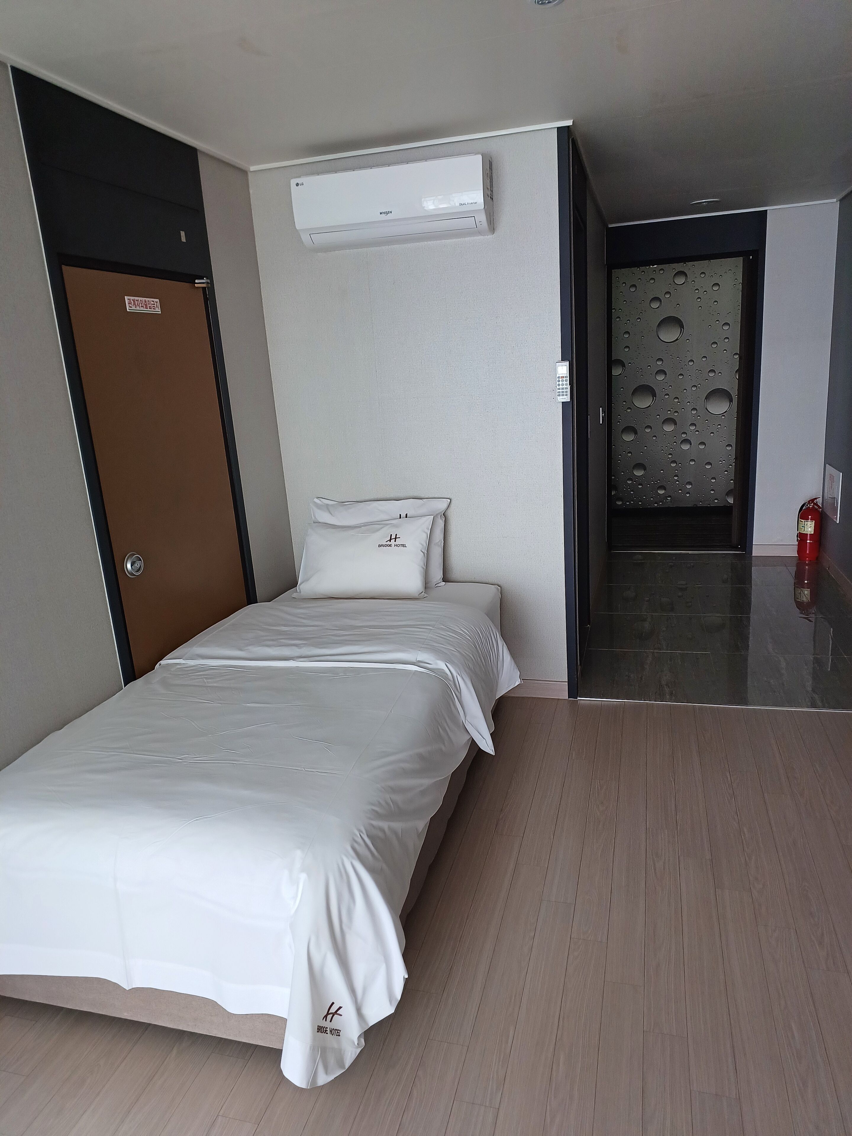 A hotel room with a bed, an air conditioning unit, a door, a wall-mounted control panel, and a fire extinguisher.