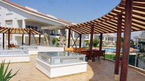 Un espacio al aire libre techado con asientos y una mesa de vidrio, rodeado por una piscina y edificios.