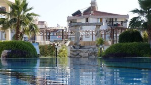 Una piscina con una estructura de piedra en el centro, rodeada de vegetación y un edificio con balcones.