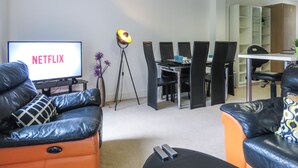 A modern living room with a TV displaying 'NETFLIX', a dining area with chairs, and a small table.