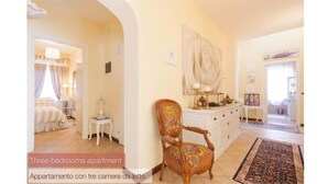 Un appartement de trois chambres à coucher, avec un couloir menant à une chambre dotée d’une grande fenêtre, d’une chaise en bois recouverte d’un tissu à motifs floraux et d’une commode blanche agrémentée d’objets décoratifs.