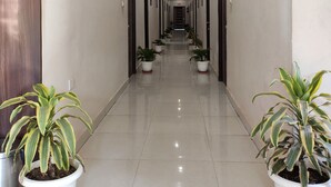 A long, narrow hotel corridor with potted plants on either side, a check-in/out sign, and a security camera. - Hotel Maple Delite (Lucknow)