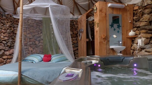 Een rustieke houten kamer met een stenen muur, een bed met hemelbed en een jacuzzi met bedieningspaneel.