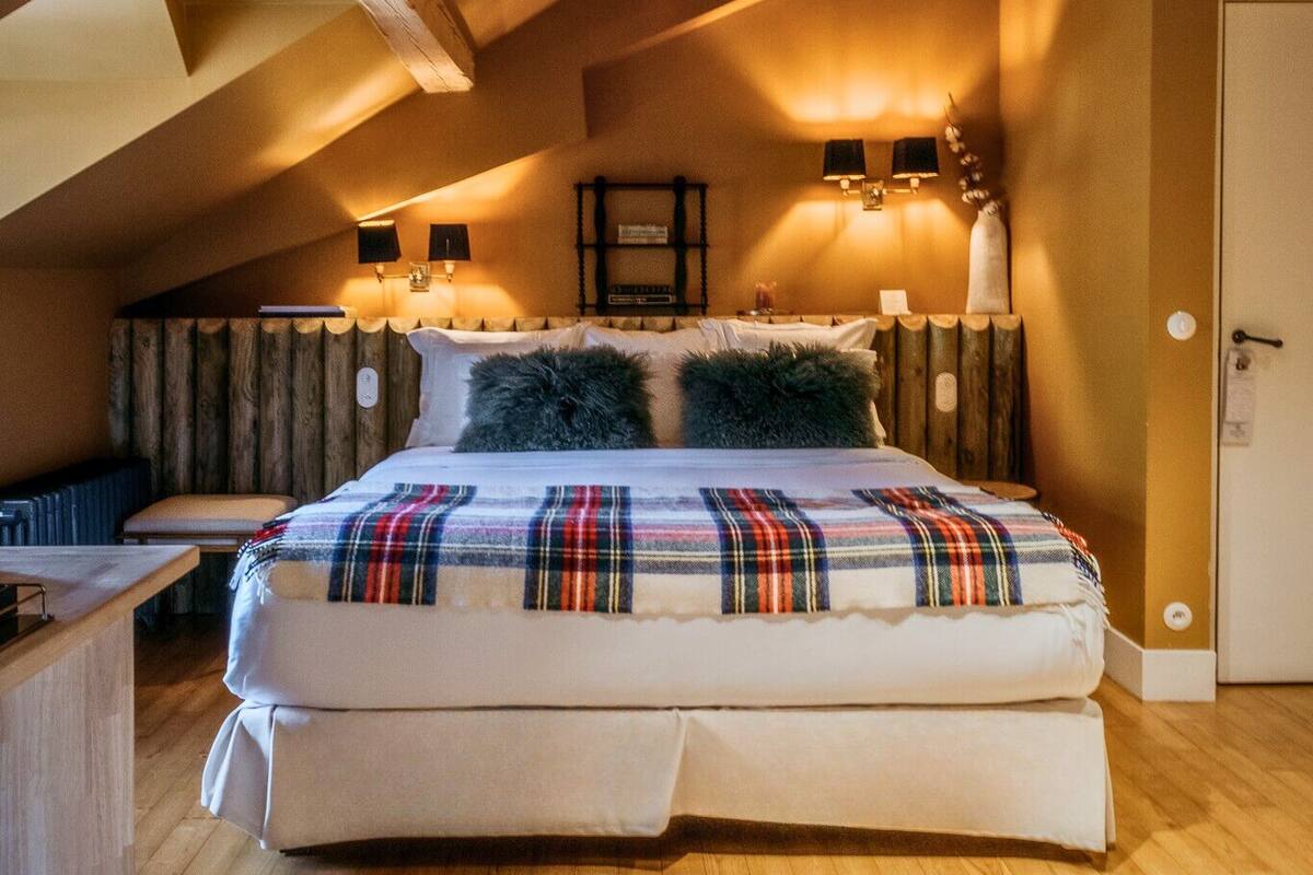 A cozy bedroom with a large bed, wooden beams, a sloped ceiling, and a small desk with a chair.