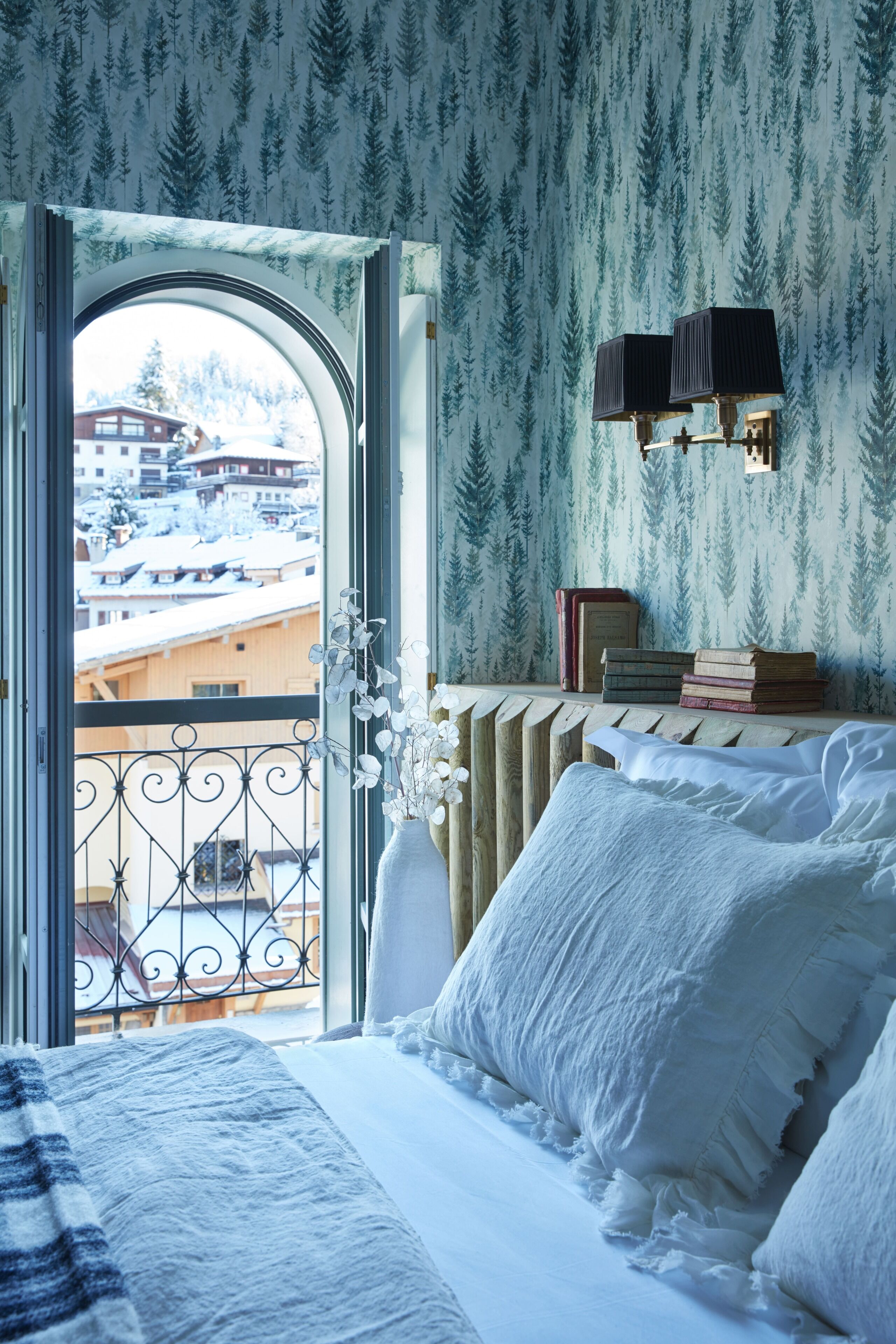 A bedroom with a bed, a radiator, a window with a view of buildings, and a shelf with books and a vase.