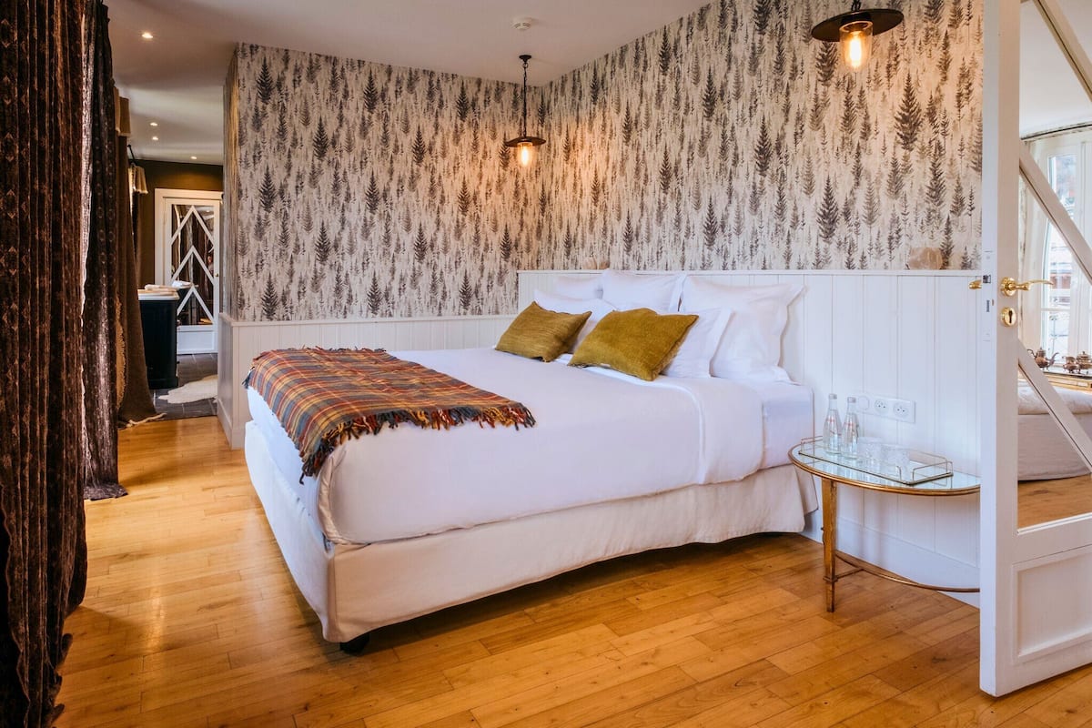 A bedroom with a large bed, two pillows, a bedside table with glasses, and a patterned wallpaper.