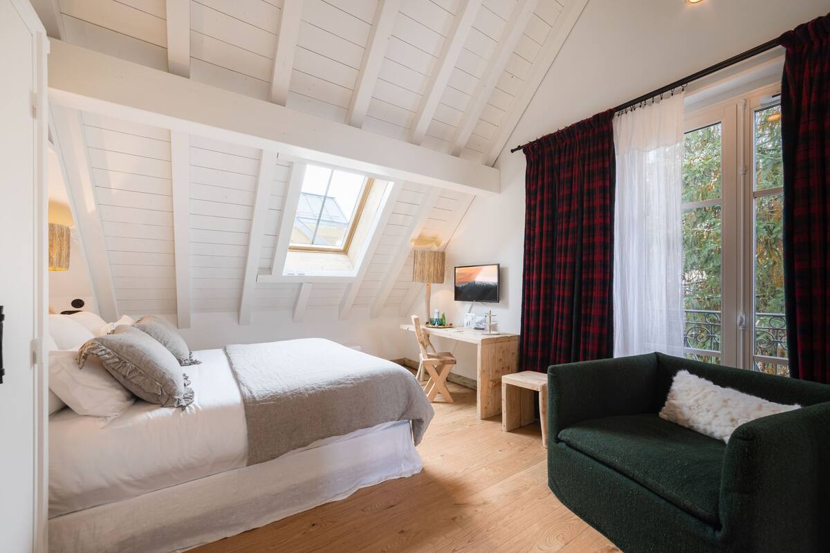 A bedroom with a bed, a desk, a chair, a couch, a TV, and a window with curtains.