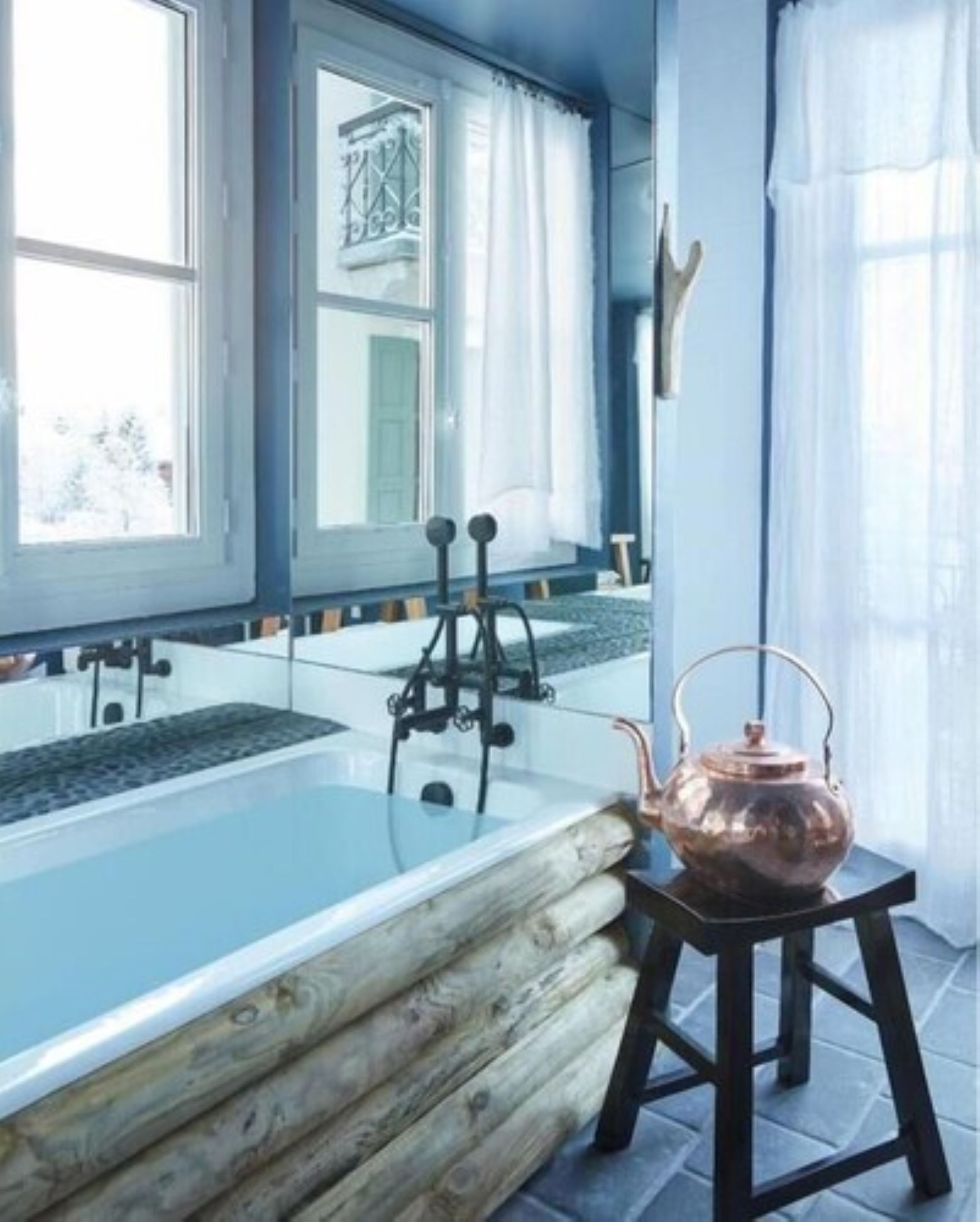 A modern bathroom with a glass bathtub, wooden log bench, and a copper kettle on a stool.