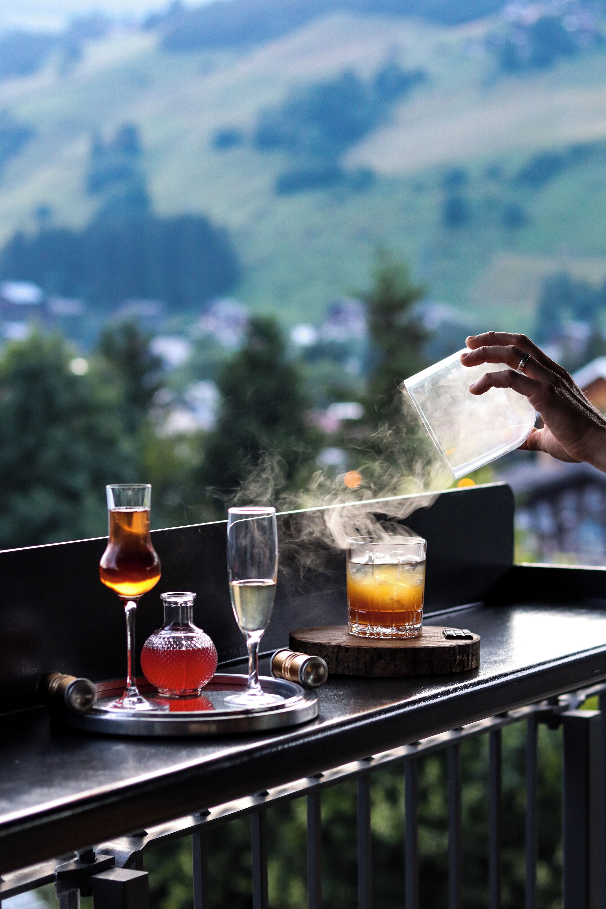 A balcony with a table holding glasses of beverages and a bottle, overlooking a scenic landscape.