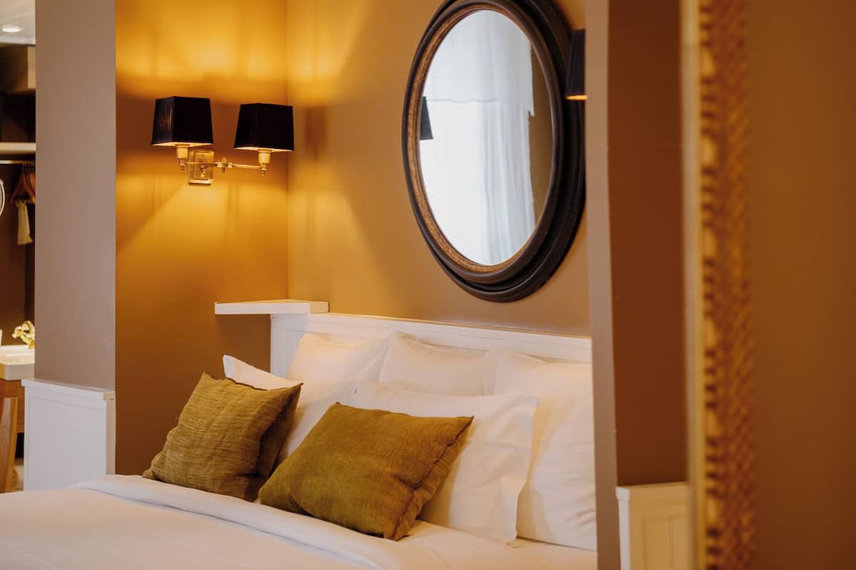 A hotel room with a bed, a round mirror, wall sconces, and a view of a hallway.