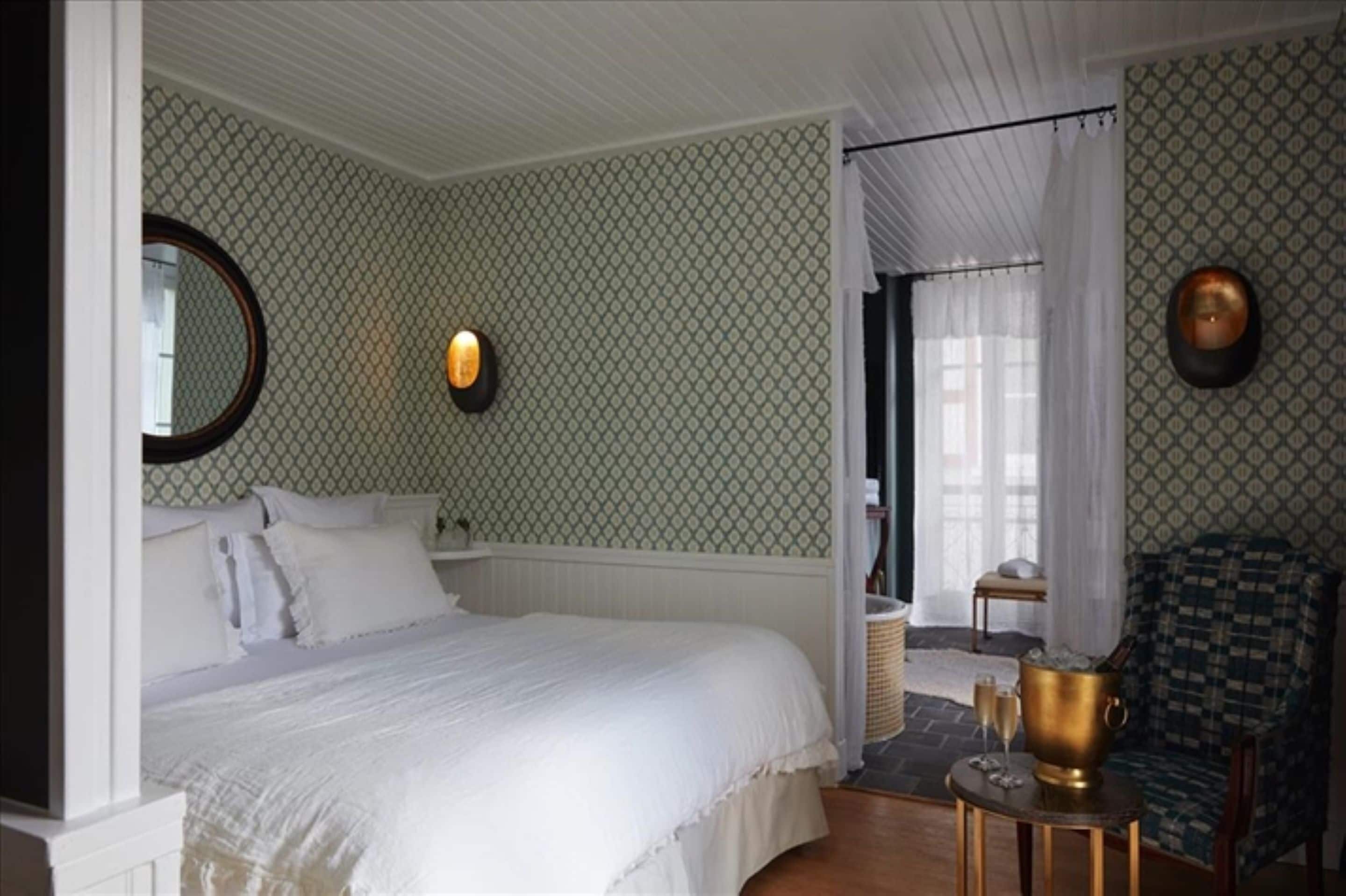A bedroom with a bed, a round mirror, a wall-mounted light, a chair, and a small table with a bucket and glasses.