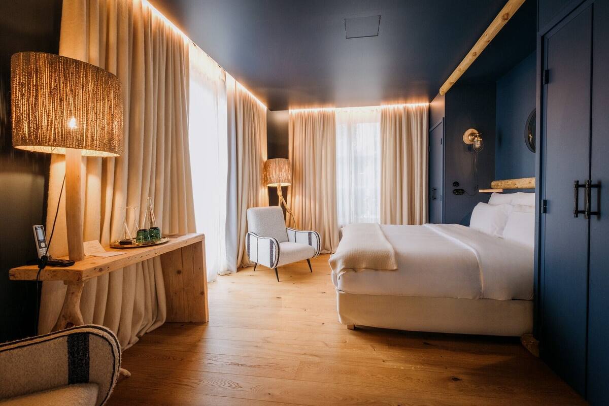A modern hotel room with a large bed, a desk with a lamp, a chair, and a wardrobe.
