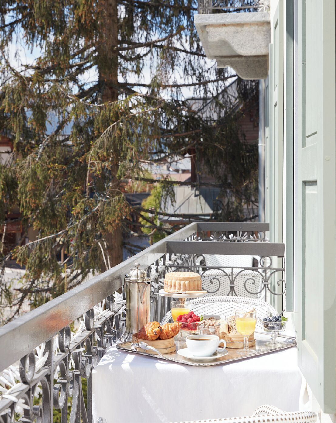 A balcony with a table set for breakfast, including croissants, a cake, and a fruit bowl.