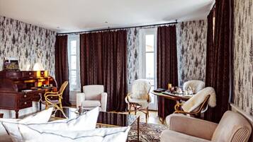 A room with a large window, dark curtains, a wooden desk, a white armchair, a beige sofa, and a patterned rug.