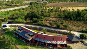 Un edificio grande de una sola planta con techo rojo y varios paneles solares. Está rodeado de campos verdes y algunos árboles dispersos.