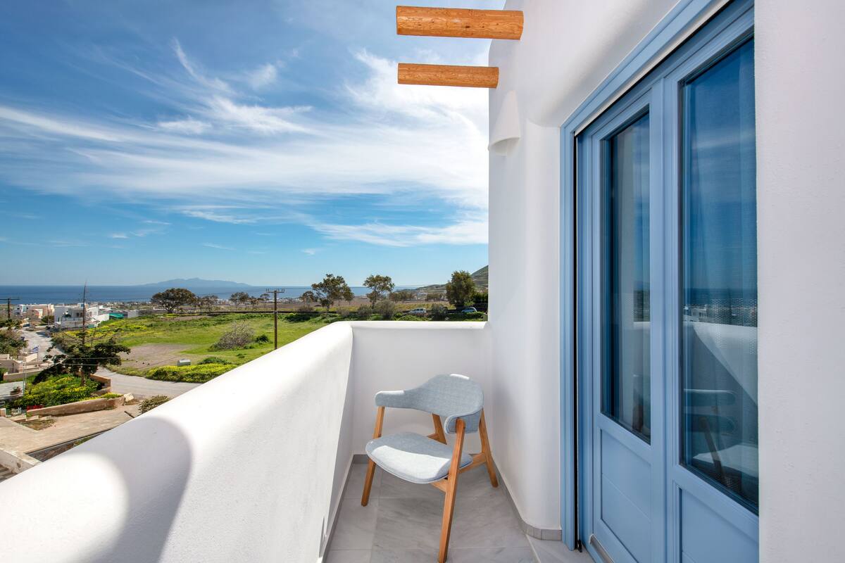 Un balcón con vistas al mar y colinas, una silla blanca y un ventanal grande.