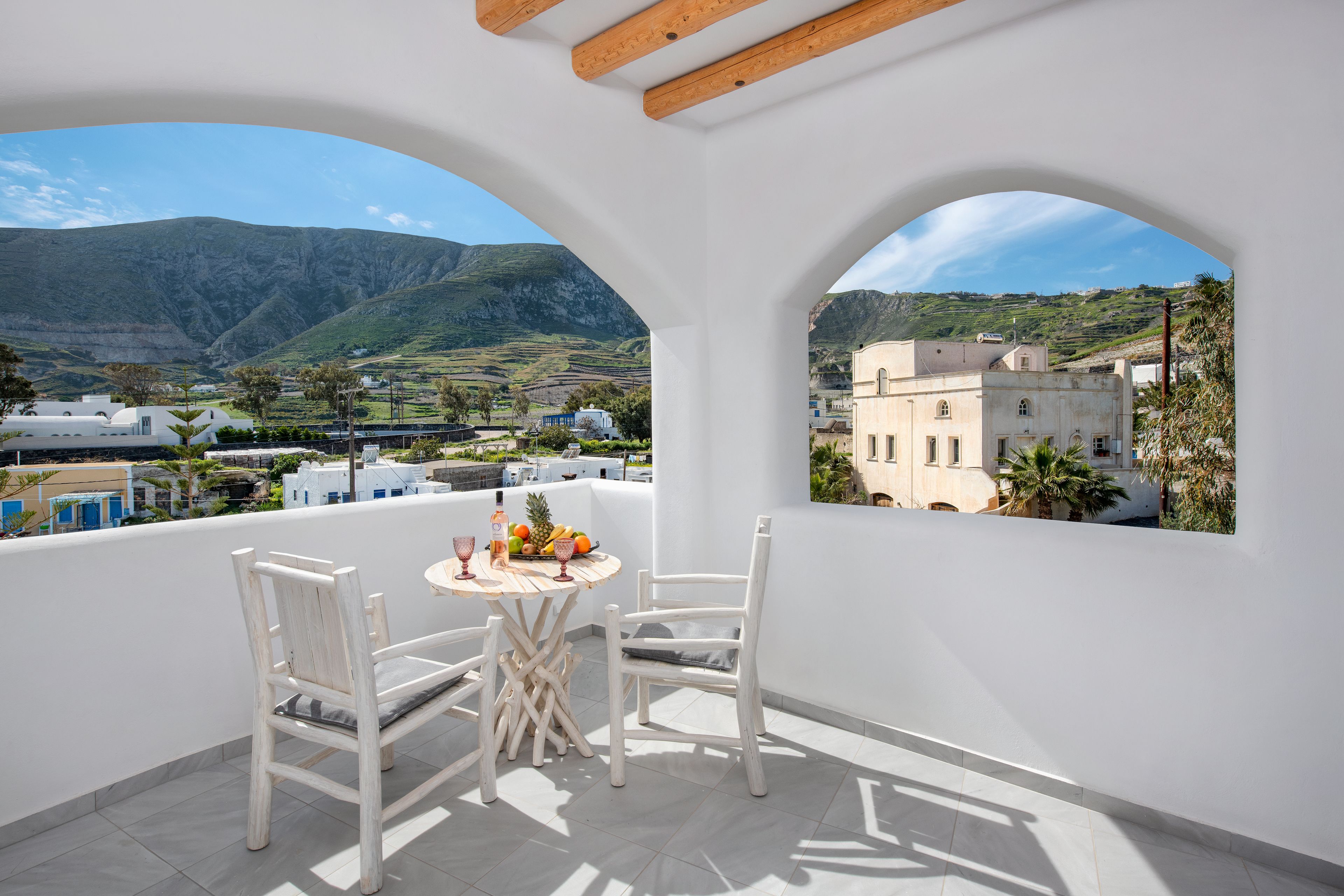 A balcony with white furniture, a table with fruits and drinks, and a view of a mountainous landscape and buildings.