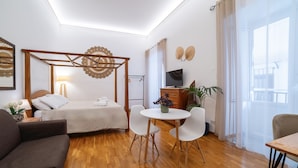 A modern hotel room with a wooden floor, a bed with white bedding, a small round table with chairs, a TV on a wooden cabinet, and a large window with sheer curtains. - Natoli Apartments - Ina House (Palermo)
