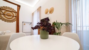 A round white table with a green vase of purple flowers, white chairs, a bed with white linens, and a wall with decorative items. - Natoli Apartments - Ina House (Palermo)