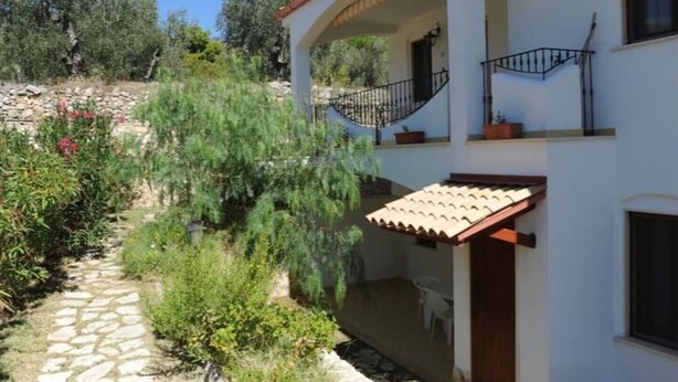 Un edificio a due piani con un portico coperto, un vialetto in pietra e vegetazione circostante.