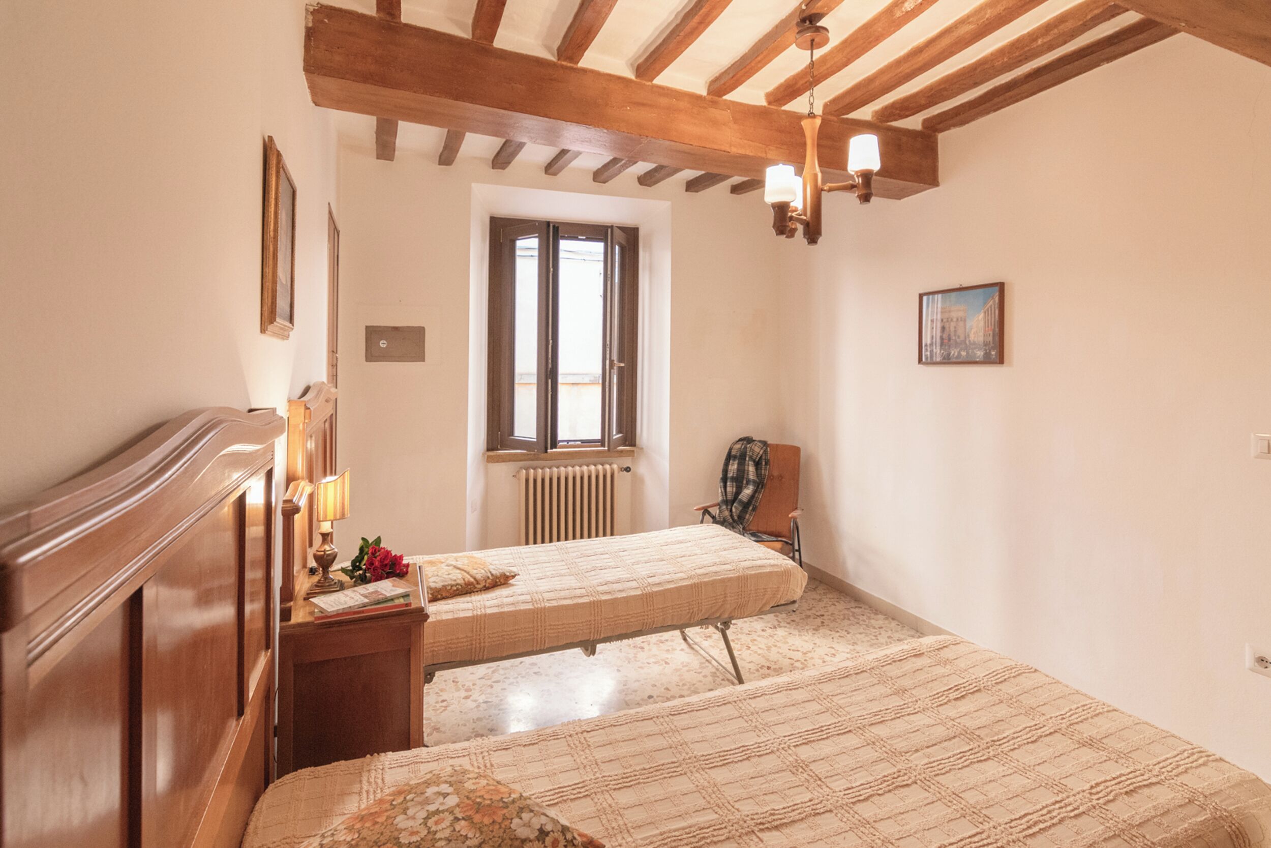 A hotel room with a wooden ceiling, a bed with a quilt, a chair, a radiator, and a window.