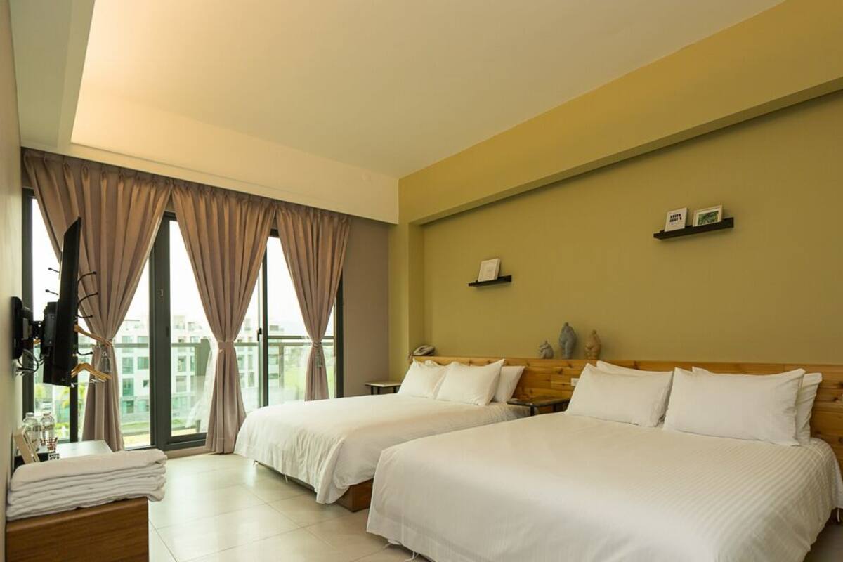 A hotel room with two beds, a wooden headboard, a large window with curtains, and a small table with a towel.