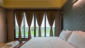 Une chambre d’hôtel avec un grand lit, une table de chevet en bois avec des serviettes, un porte-manteau et une vue sur un paysage verdoyant, visible à travers de grandes fenêtres équipées de rideaux.