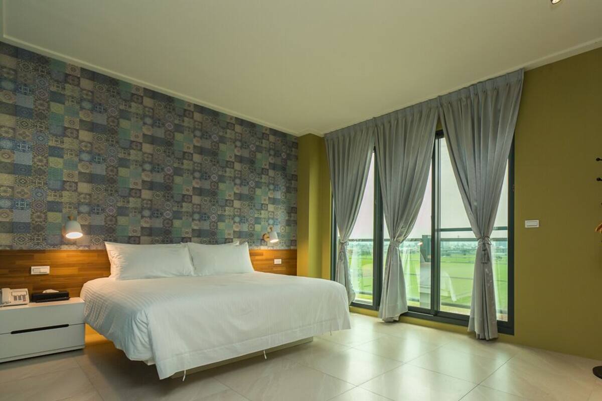 A bedroom with a large bed, a nightstand, a lamp, a coat rack, and a view of a green landscape through the window.
