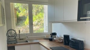 Et lille køkken med hvide skabe, en træbordplade, en mikrobølgeovn, en brødrister og et vindue med udsigt til grønne områder.