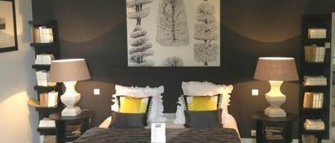 Un dormitorio con una cama, dos mesitas de noche, dos lámparas y un cuadro colgado en la pared con un paisaje de árboles.