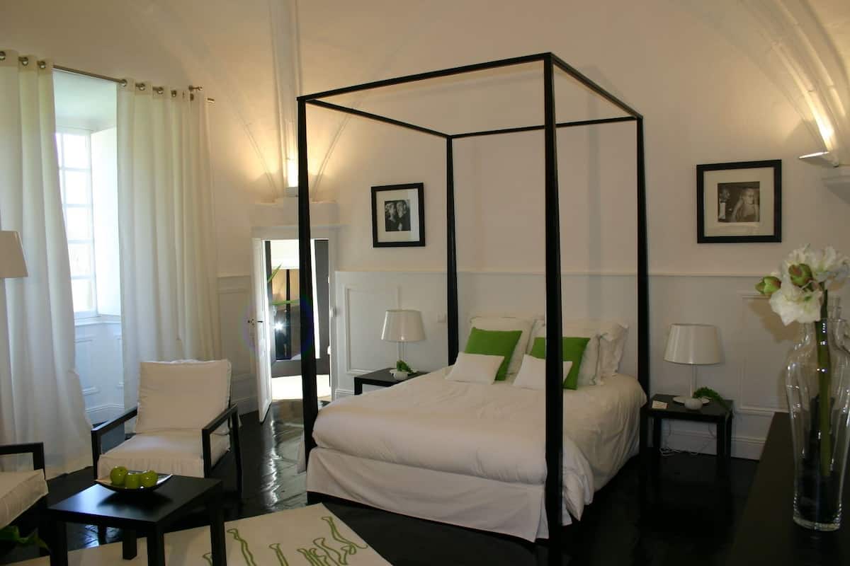 A four-poster bed with white linens and green pillows, a bedside table with a lamp, and a vase with flowers on the nightstand.