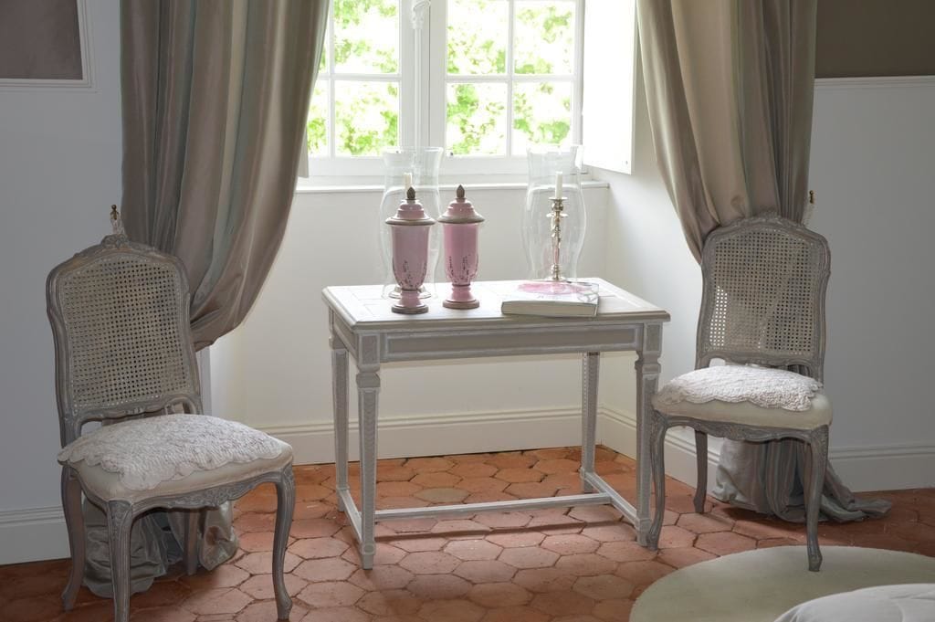 Een kamer met twee rieten stoelen, een witte tafel met een boek en kandelaars, en een raam met gordijnen.