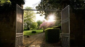 Um portão de ferro forjado aberto que dá para um jardim com um sebede e um caminho banhado pelo sol.