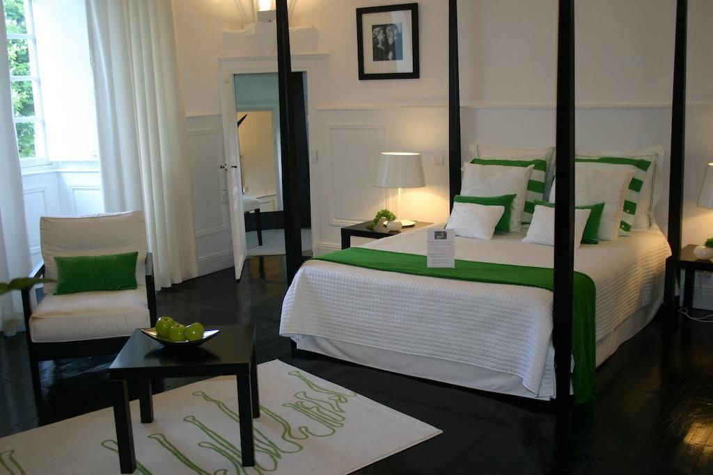 A four-poster bed with white bedding and green accents, a bedside table with a lamp, a chair with a green pillow, and a framed picture on the wall.