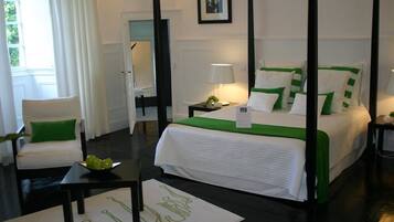 A four-poster bed with white bedding and green accents, a bedside table with a lamp, a chair with a green pillow, and a framed picture on the wall.