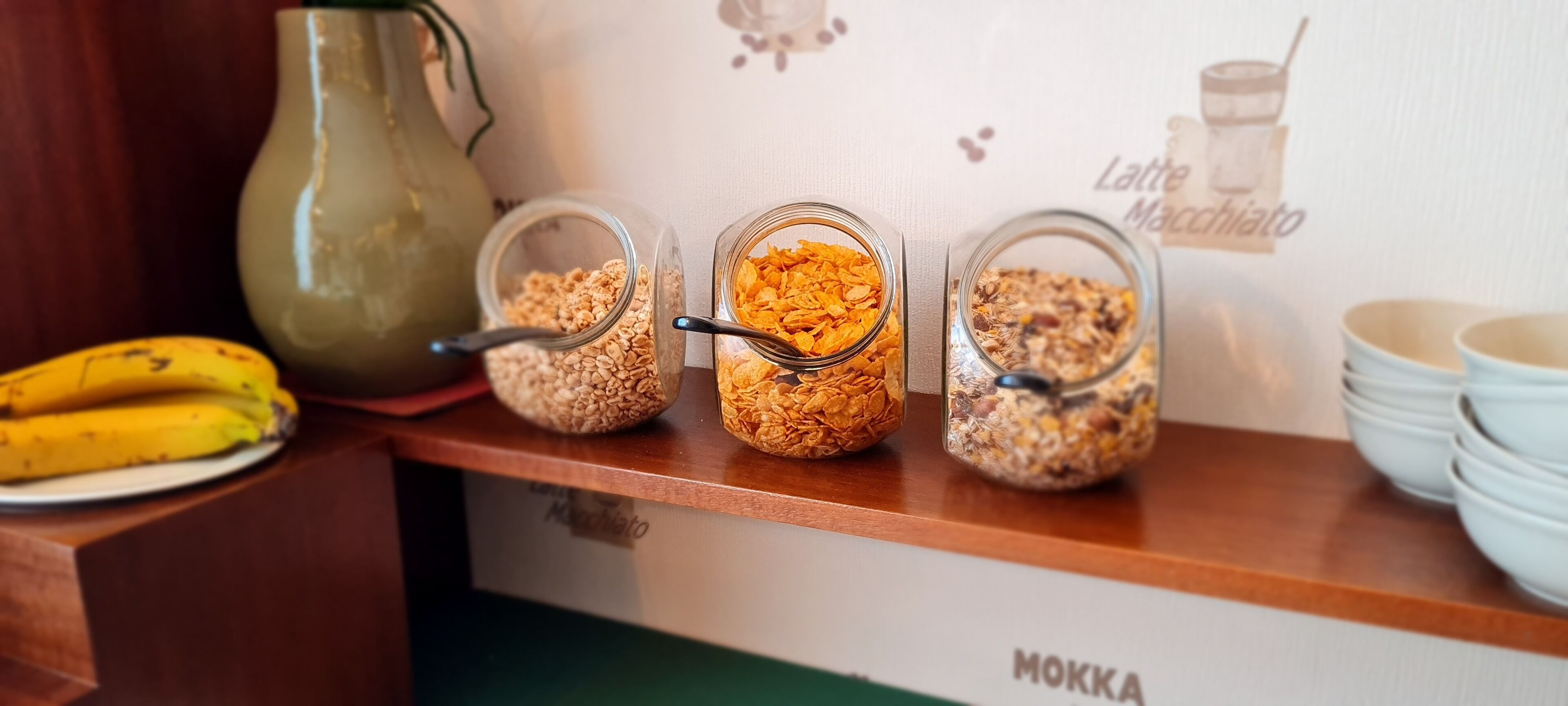 A breakfast bar with three glass containers of cereal, a banana, and a green pitcher.
