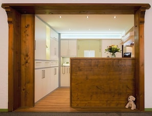 A narrow hallway leading to a kitchen with wooden cabinets, a wooden reception desk, and a small plush toy on the floor. - Hotel Garni Försterhof (Kronach)