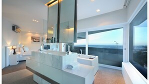 A modern bathroom with a large mirror, dual sinks, and a bathtub. It features a large window with a view of the ocean. - Ebb Tide (Cape Town)